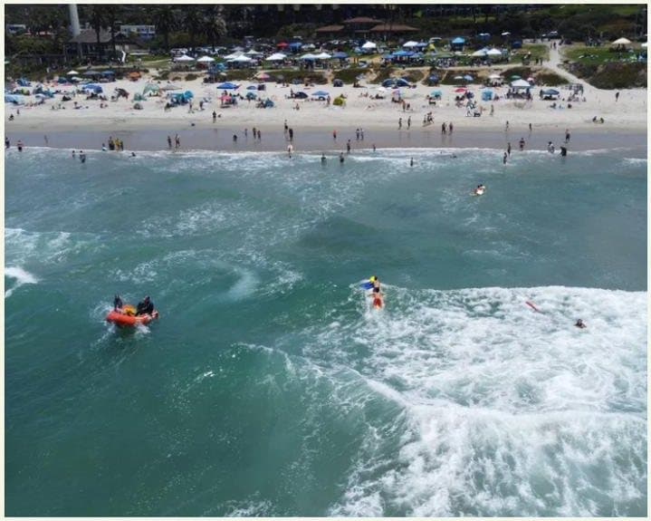 Aerial view of Del Mar lifeguards on rescue boards and an inflatable rescue boat working the surf just off a crowded beach.