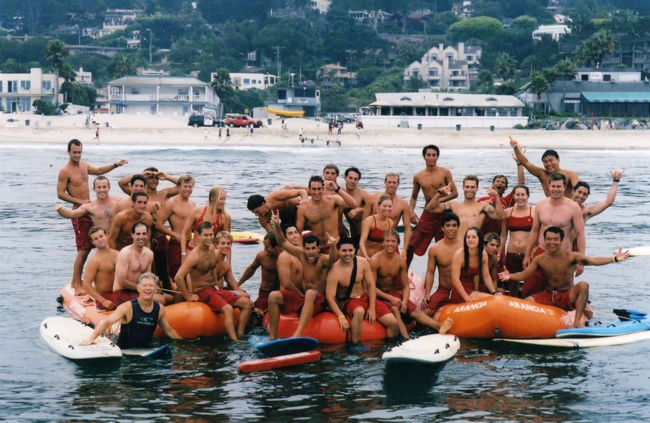 The 2007 Del Mar lifeguard crew gathered on rescue boats and surfboards in the ocean, with the beach behind them.