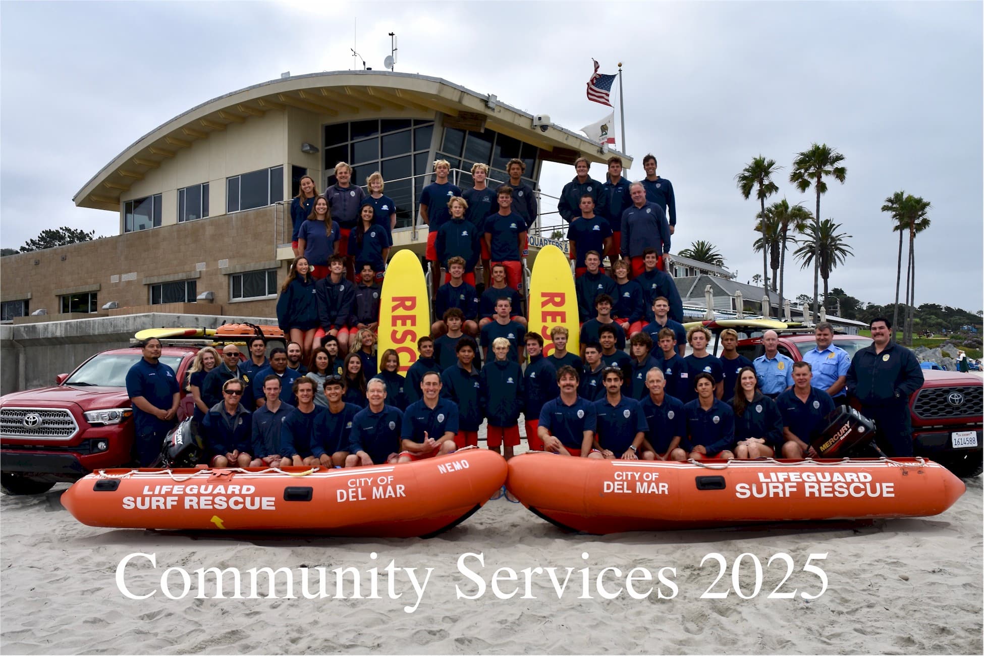 2025 Del Mar lifeguard team photographed on the sand in front of lifeguard headquarters with two rescue boats.